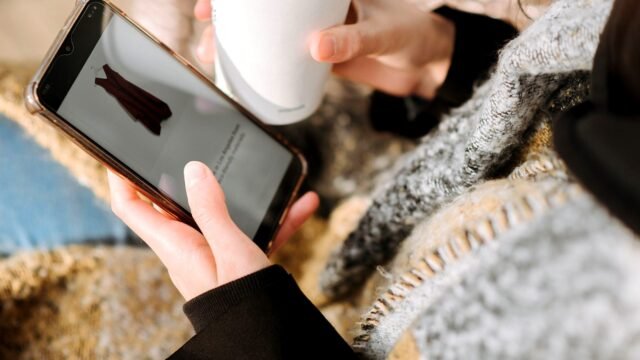 A shopper holds a phone showing a dress in an online shop, with a coffee cup beside it