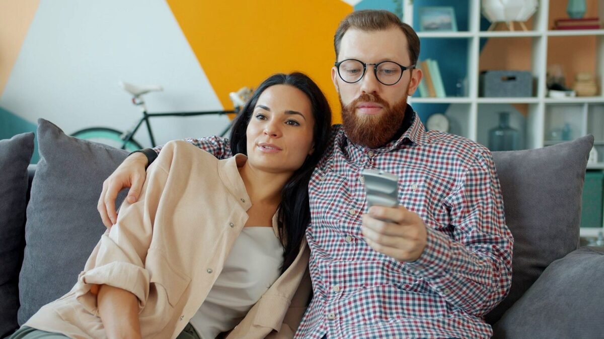 Two people sit on a sofa at home watching a television across the room