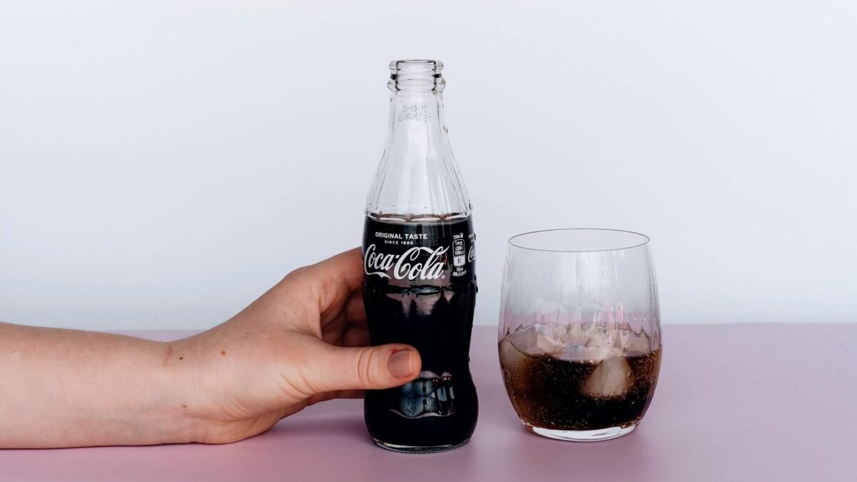 A hand holds a glass bottle of Coca-Cola next to a filled glass on a light table.
