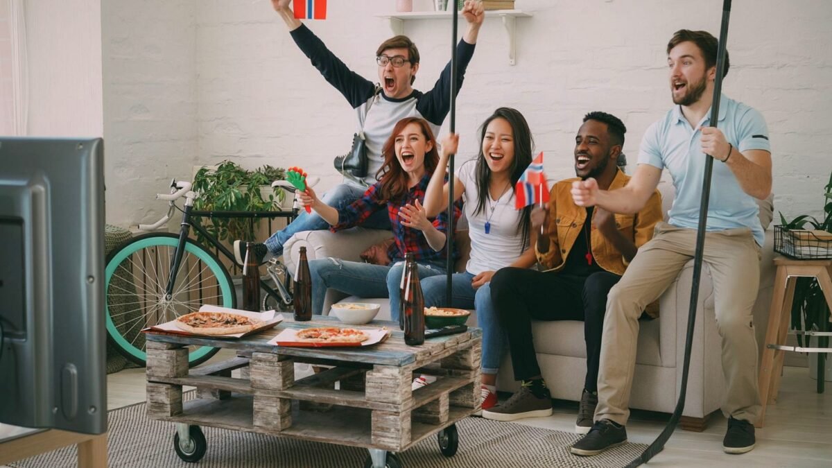 Excited friends cheering while watching a sports game on TV, holding flags and snacks.