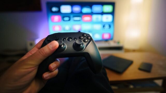 Close-up of a person holding a gaming controller in a cozy living room setup with TV.