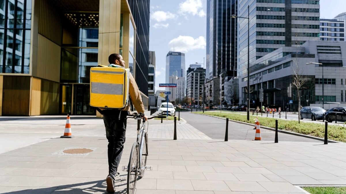 A bicycle courier with a thermal delivery bag rides along a city street between cars.
