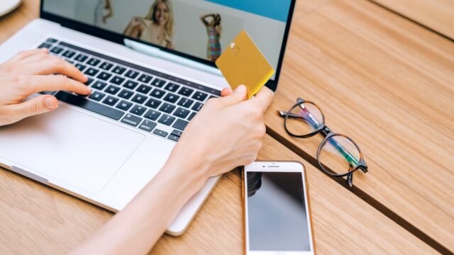 A person makes an online purchase using a credit card on a laptop, with a smartphone and glasses nearby.