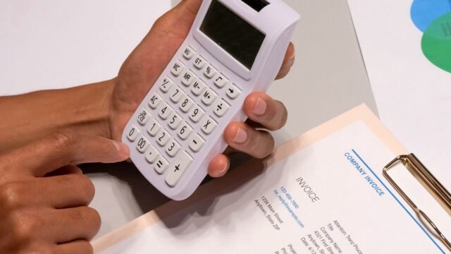 Hands work a calculator beside printed invoices on an office desk.