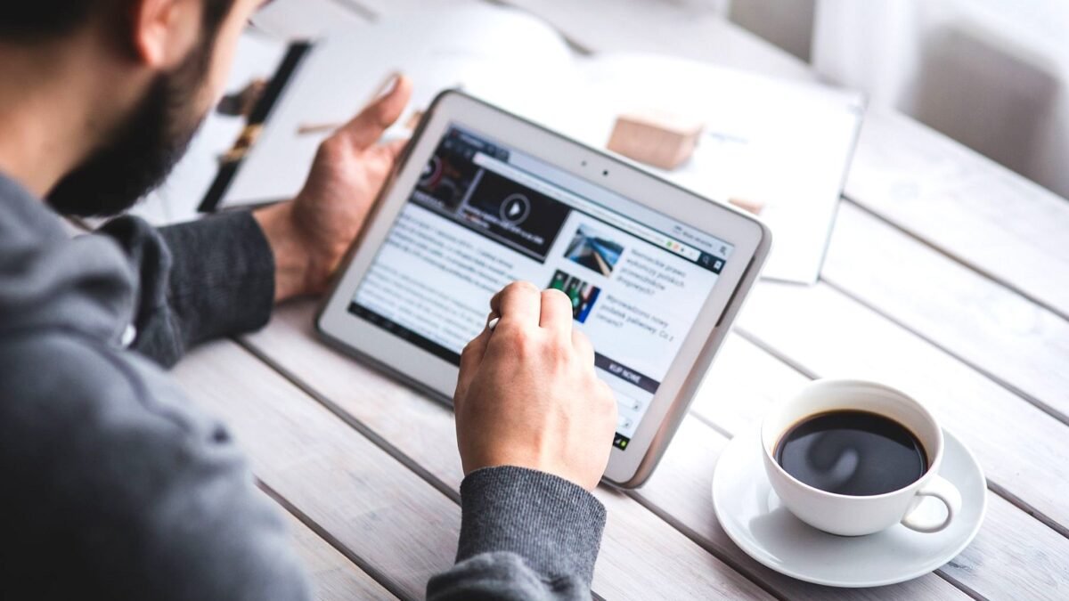 A man reads news on a tablet at a table with a cup of coffee beside him.