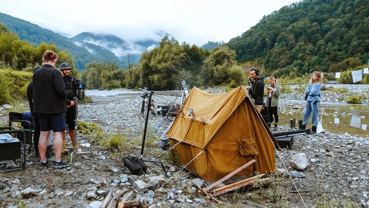 A small film crew sets up cameras and a tent by a river in a wooded valley