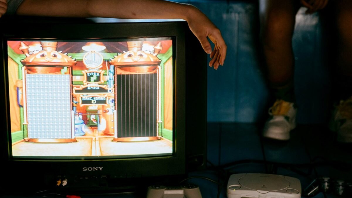 A young man on a sofa watches gameplay footage on a large TV with a game controller resting beside him.