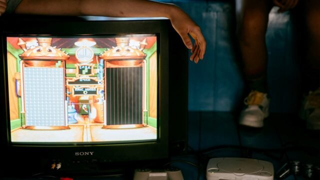 A young man on a sofa watches gameplay footage on a large TV with a game controller resting beside him.