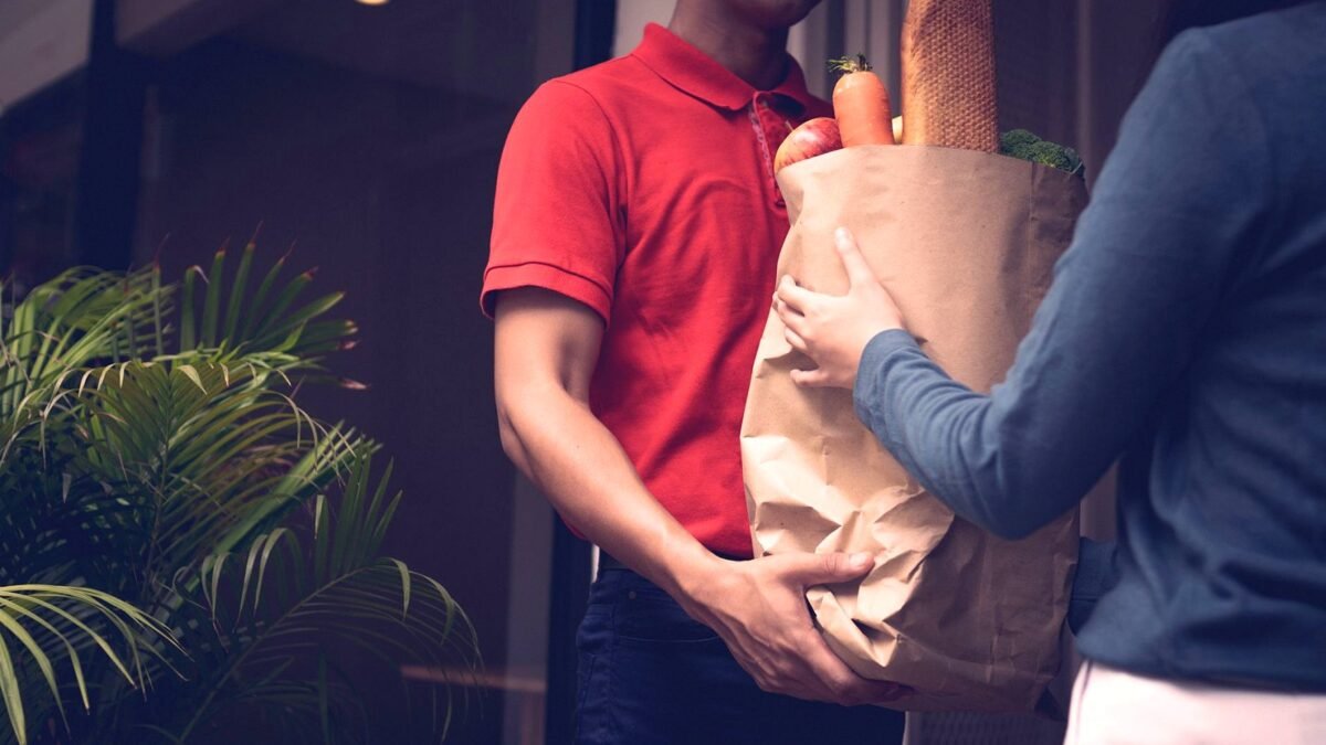 A delivery worker carries a bag of groceries to a customer at the door.