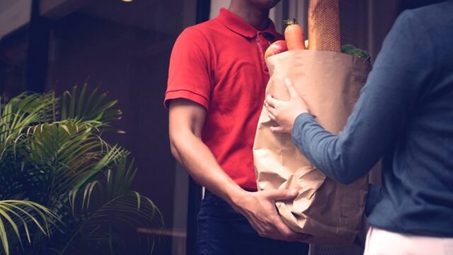 A delivery worker carries a bag of groceries to a customer at the door.