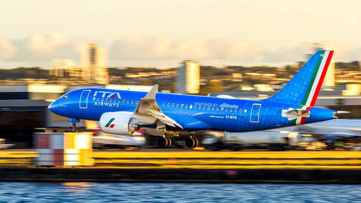 An ITA Airways jet climbs away from the runway with a city skyline behind it