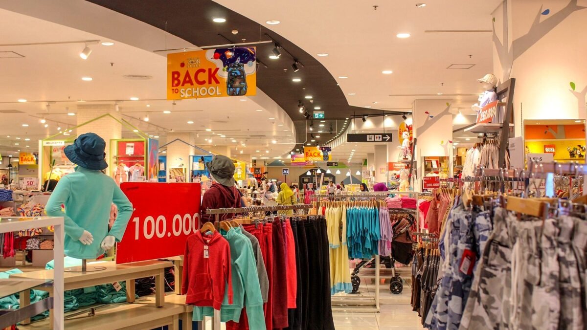 A bright department store aisle with clothing rails and shoppers walking past.