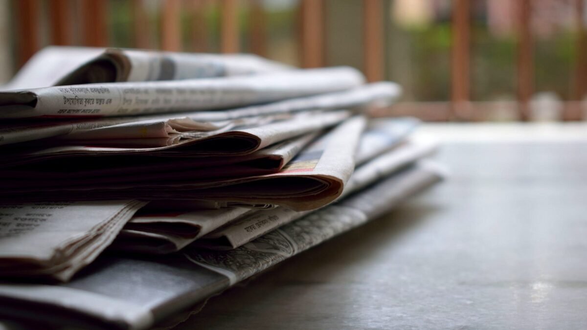 A newspaper newsroom with journalists at desks and monitors, printed pages in the foreground.