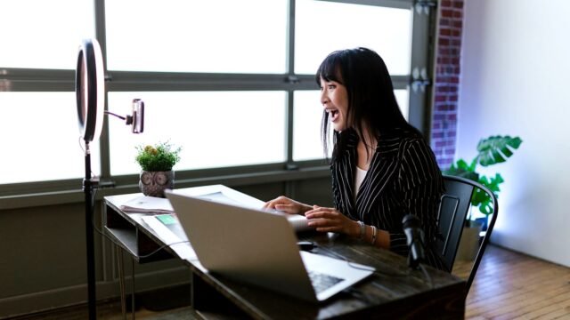A woman records a video at an office desk with a laptop and a small camera in front of her.
