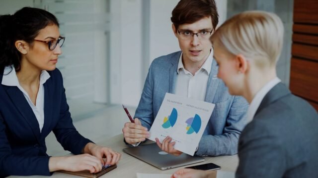 Several people around a table look at sales charts on a screen in a bright office.