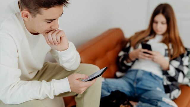 Two teenagers sit on a sofa, each looking at the screen of a smartphone.