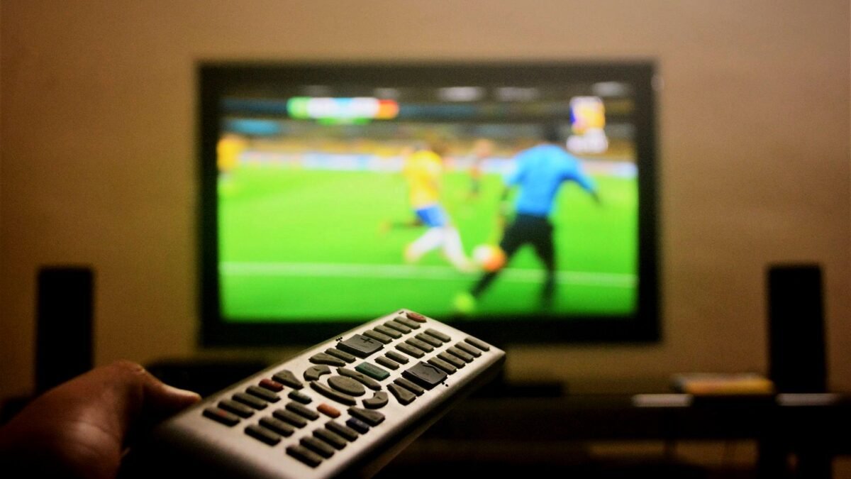 Hand with a remote control in front of a TV showing a football match