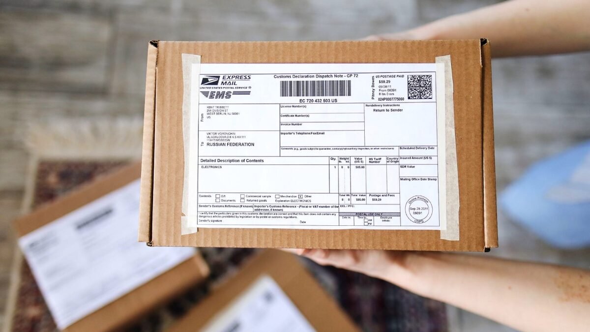 Hands holding a small cardboard parcel with a customs declaration form stuck to the top.