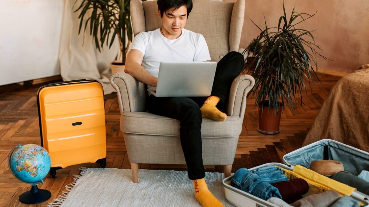 Man sitting on armchair planning travel, using laptop, with suitcases and globe nearby.