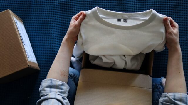 Hands open a cardboard parcel with a folded white T-shirt inside, on a table at home.
