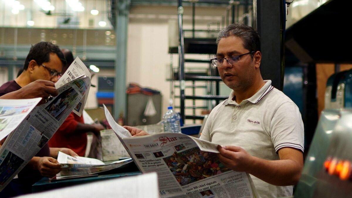 Workers at a newspaper printing press reading freshly printed copies. Focus on a man holding a newspaper.