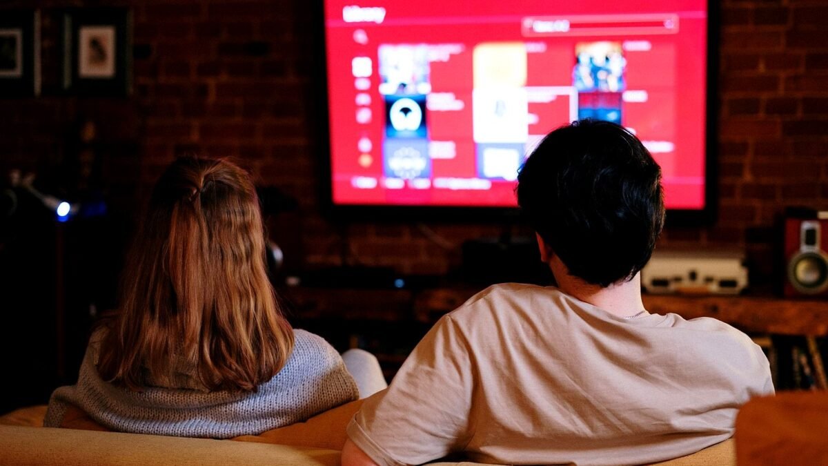 Two people sit on a sofa in front of a large television showing a film