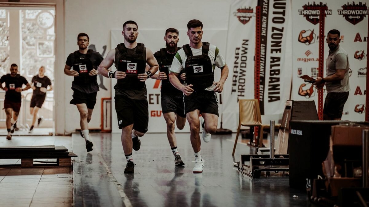 Athletes running side by side in an indoor fitness race hall watched by a crowd.