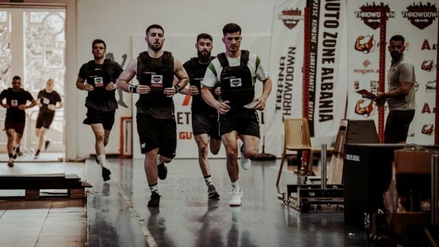 Athletes running side by side in an indoor fitness race hall watched by a crowd.