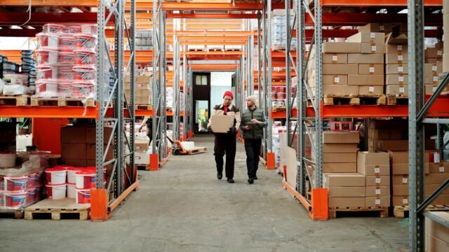 Two workers handle a package in a spacious warehouse surrounded by shelves stocked with boxes and products.