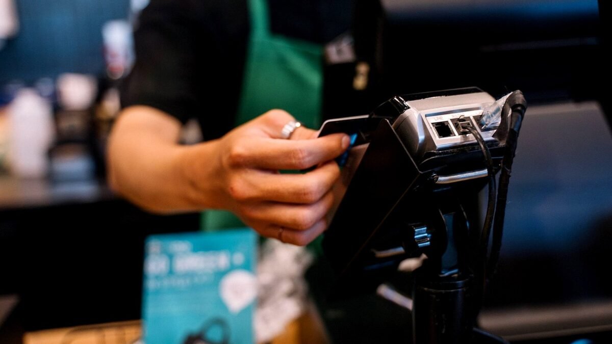 A barista holds a card terminal across a cafe counter while a customer pays