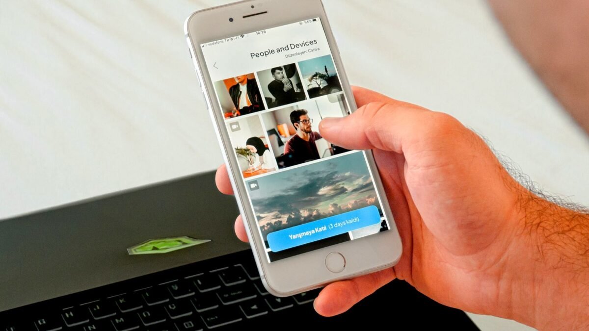 A hand holds a phone and swipes through photos, with a laptop open behind it