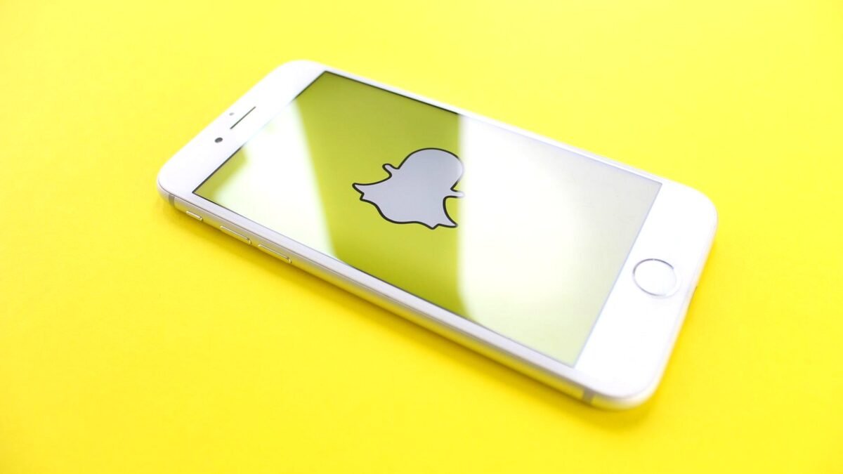A white phone lying on a bright yellow surface, its screen showing the Snapchat logo.