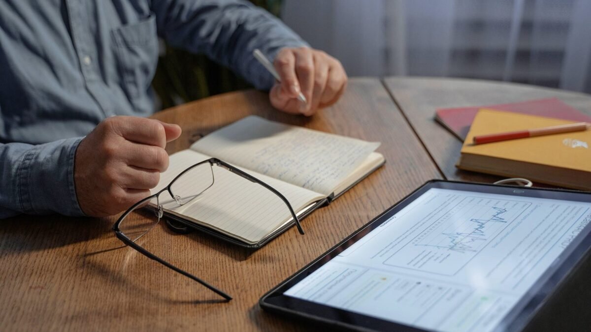 Man making notes next to a tablet showing a chart on a wooden desk