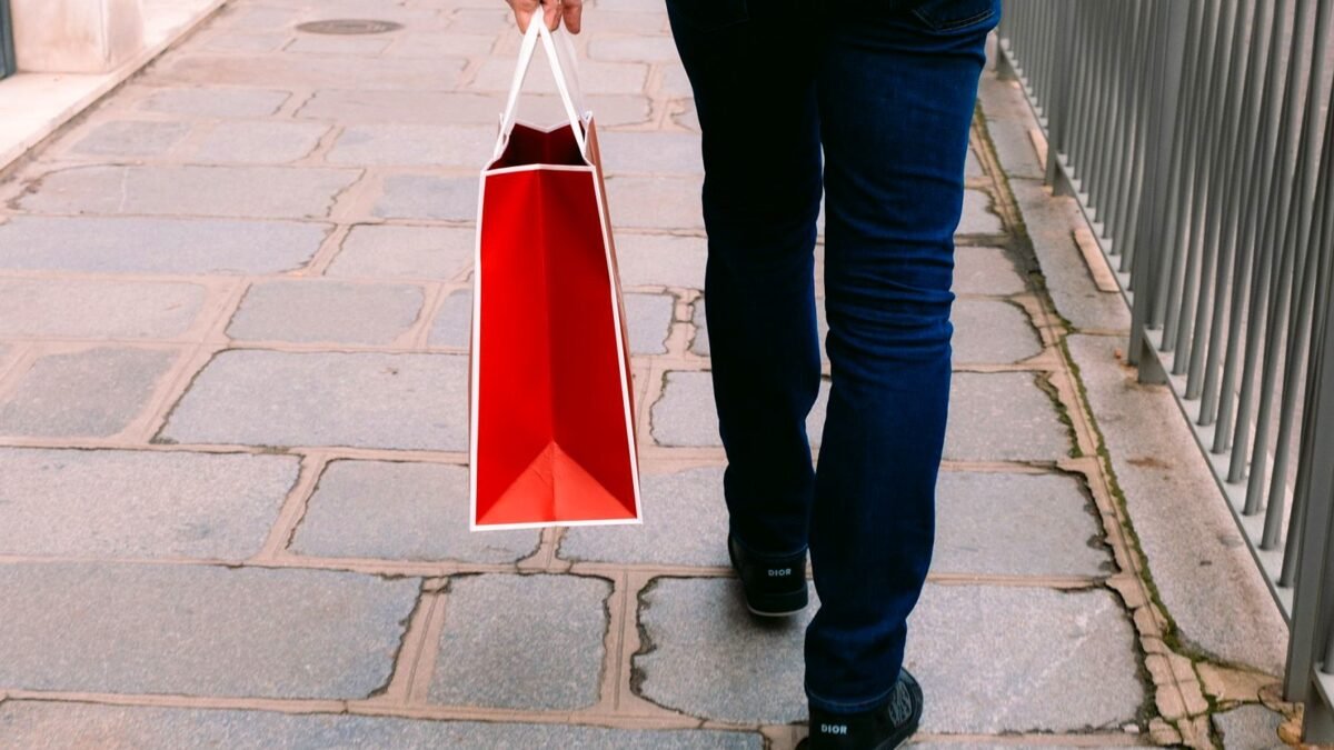 A shopper walks along a street carrying a red paper shopping bag.