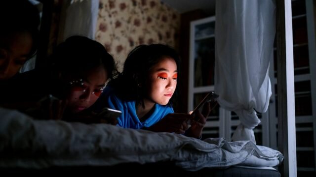 Three teenagers sit together in a bedroom, each looking at a smartphone.