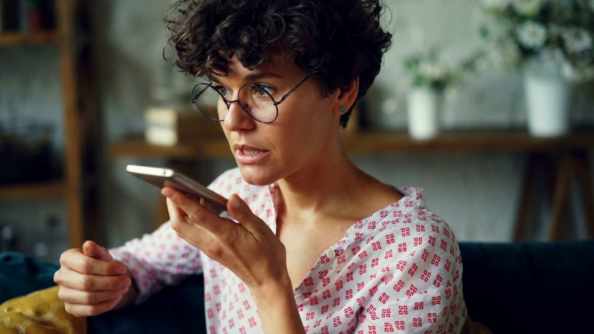 A woman sitting at home speaks into the smartphone she holds close to her face.
