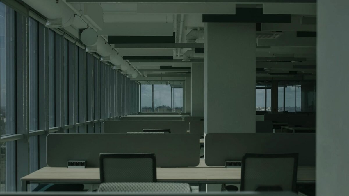 Rows of empty desks in a bright open-plan office with large windows