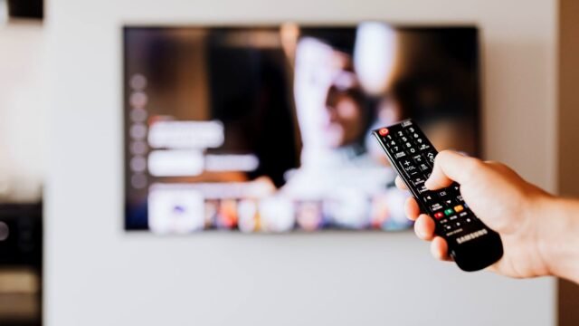 Hand holding a TV remote control in front of a blurred television screen