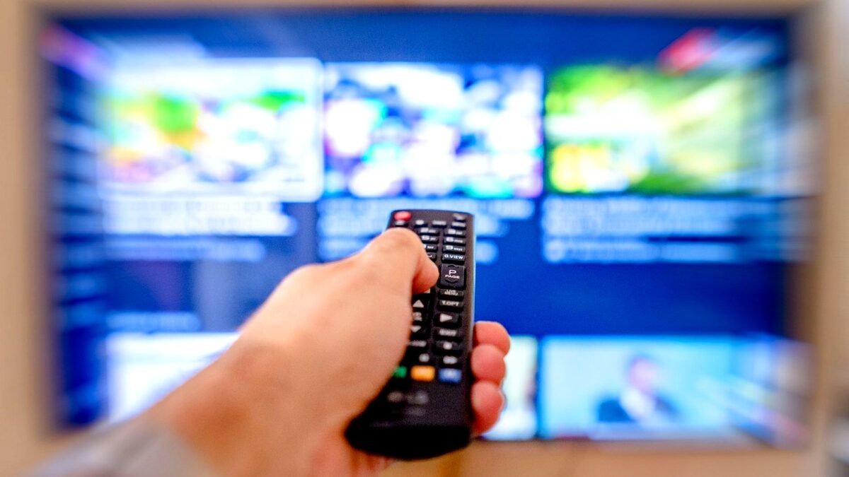 Hand holding remote control pointing at a blurred TV screen in a cozy indoor setting.