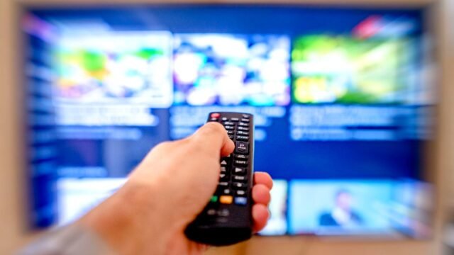 Hand holding remote control pointing at a blurred TV screen in a cozy indoor setting.