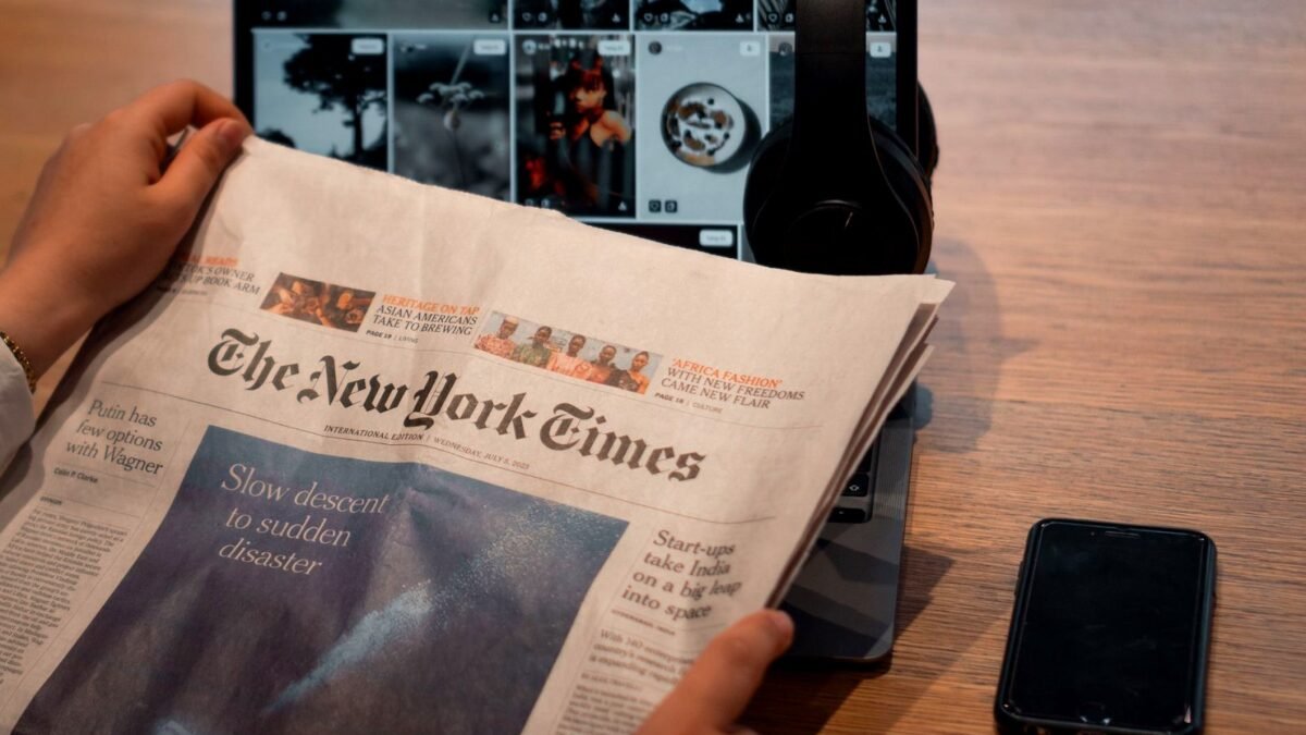 Hands holding a printed newspaper beside a laptop and a phone on a table