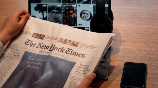 Hands holding a printed newspaper beside a laptop and a phone on a table
