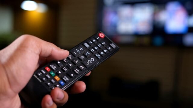 A hand holding a TV remote control in front of a blurred television screen