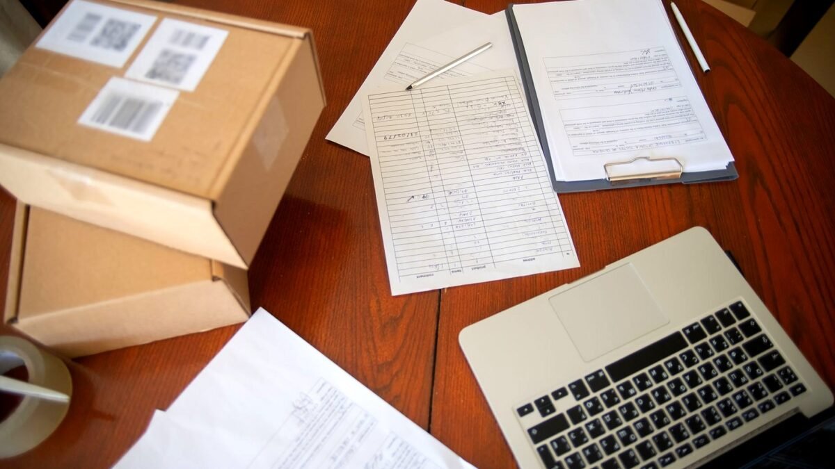 Laptop, documents and cardboard shipping boxes on a desk, seen from above.