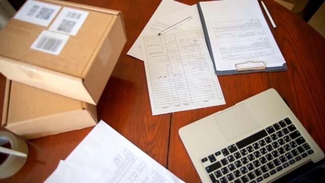 Laptop, documents and cardboard shipping boxes on a desk, seen from above.