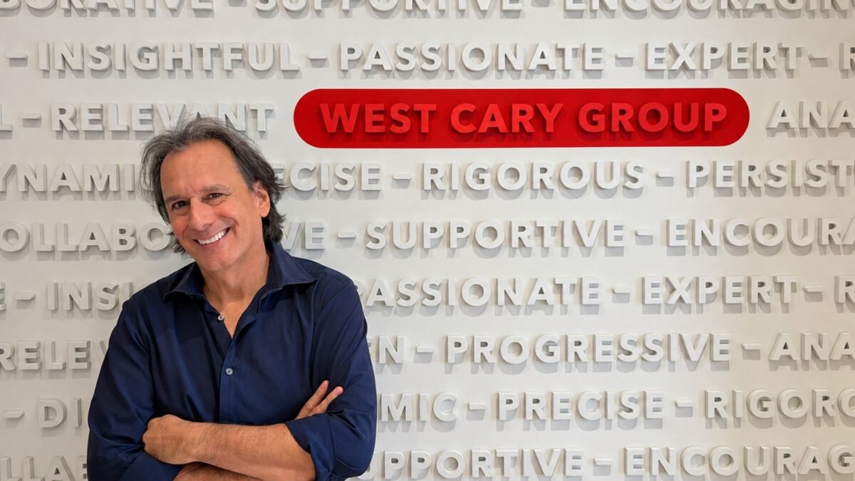 Andy Azula in a navy shirt, arms crossed, in front of a wall of West Cary Group values with the agency logo in red