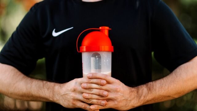 Hand holding a protein shaker with a red lid outdoors