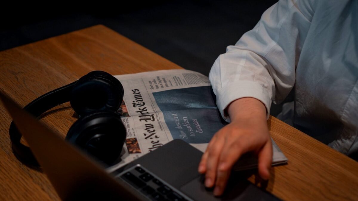 Person with headphones working on a laptop next to a printed newspaper