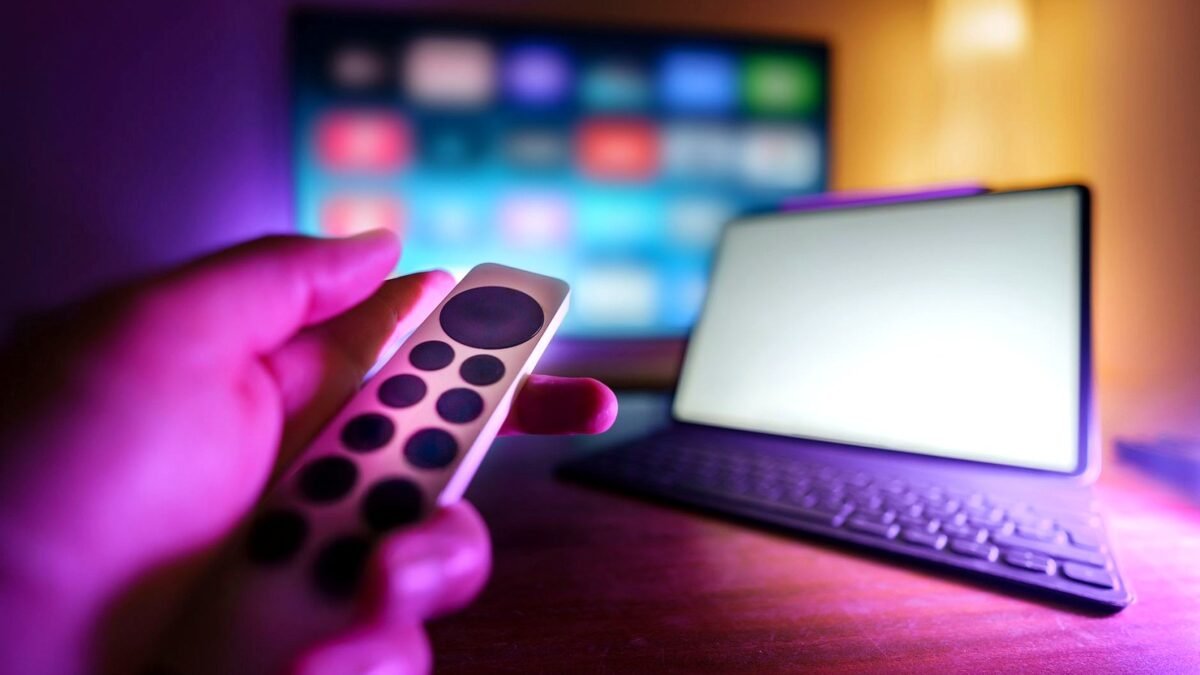 Hand using a remote to control a TV next to a laptop