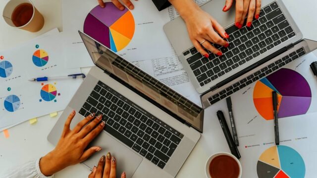 Two people working on laptops over printed pie and bar charts, seen from above.